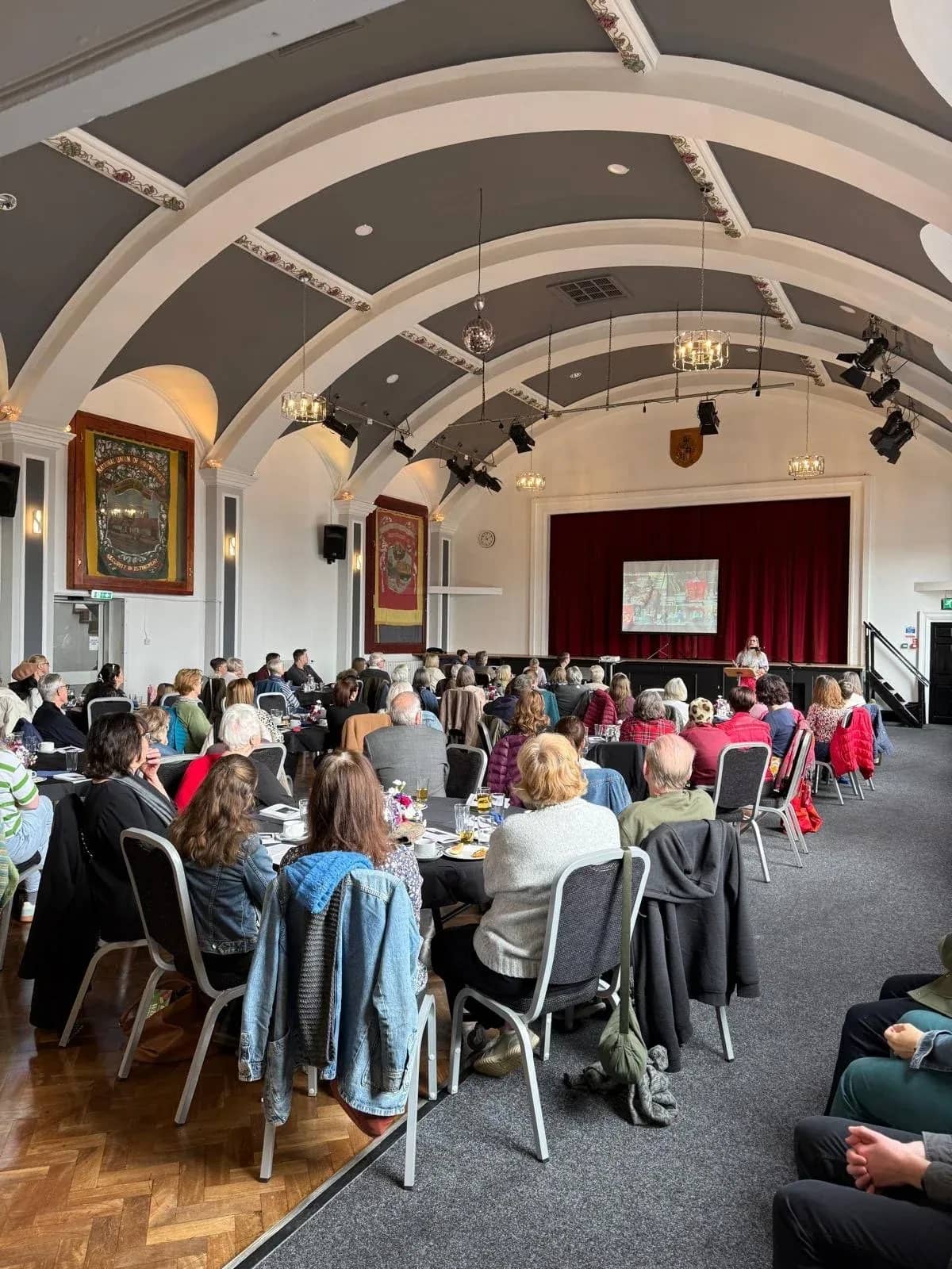 A full room listening to a talk under an arched ceiling